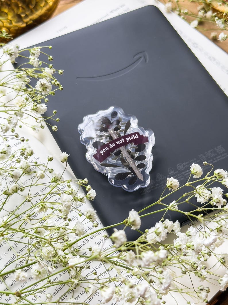 May include: A black e-reader with a clear resin embellishment. The resin piece contains dried flowers and a burgundy ribbon with the text "you do not yield." White flowers and a book are in the background.