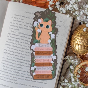 Puede incluir: Un marcapáginas caprichoso con un gato naranja sosteniendo una flor, posado sobre una pila de libros con títulos humorísticos. El marcapáginas está colocado sobre un libro abierto con detalles florales, creando un tema literario encantador.