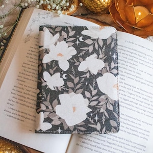 May include: A black book cover with a floral design featuring white magnolia flowers and leaves. The background is a dark gray with white stars. The book cover is made of a soft, textured material.