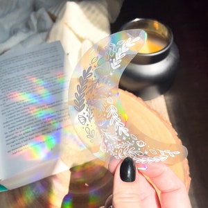 May include: A holographic crescent moon sticker with a white floral design. The sticker is iridescent and has a rainbow sheen.