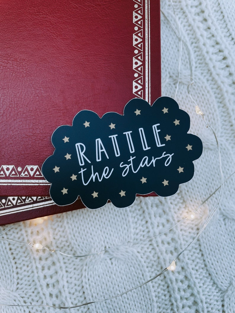 Peut inclure: Un autocollant noir en forme de nuage avec des &eacute;toiles dor&eacute;es et le texte "RATTLE the stars" sur une surface en tricot blanc avec un livre rouge en arri&egrave;re-plan.