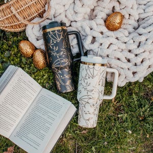 May include: Two stainless steel travel mugs with black and white bat designs and gold accents. The mugs are on a green grassy surface with a book and a white blanket.