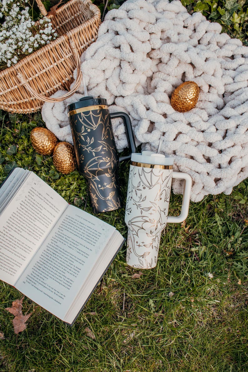 May include: Two stainless steel travel mugs with black and white bat designs and gold accents. The mugs are on a green grassy surface with a book and a white blanket.