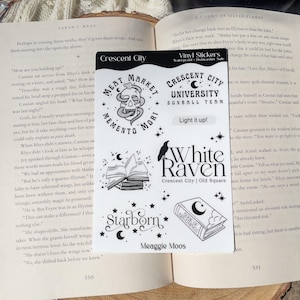 May include: A collection of white vinyl stickers with black illustrations and text. Designs include a skull with a snake, open books, stars, a raven, and the text "Crescent City" and "White Raven". The stickers are on a book.
