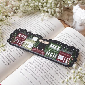 May include: A black and white illustrated bookmark featuring a row of whimsical shops with festive Christmas decorations. The shops include "Santa's Workshop", "The Gingerbread Bakery", "Hot Cocoa", "The Wishing Well", and "North Pole Candy", with a snowy background.