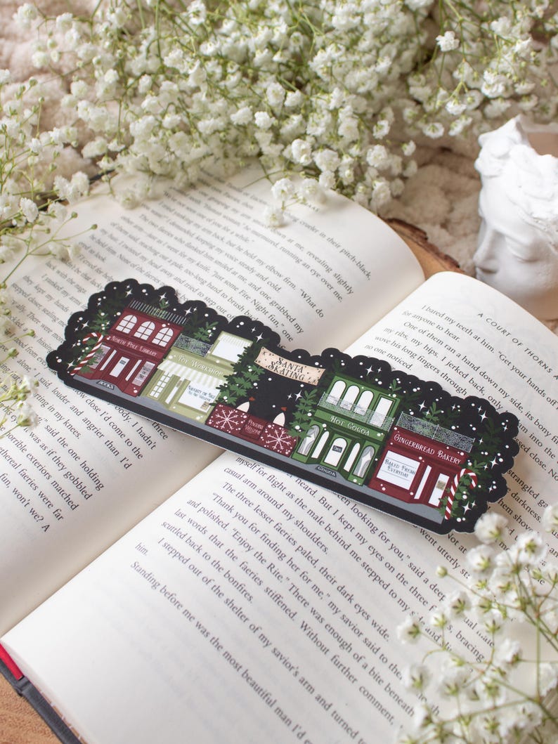 May include: A black and white illustrated bookmark featuring a row of whimsical shops with festive Christmas decorations. The shops include "Santa's Workshop", "The Gingerbread Bakery", "Hot Cocoa", "The Wishing Well", and "North Pole Candy", with a snowy background.