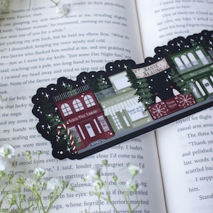May include: A black and white illustrated sticker featuring a winter village scene with five buildings. The buildings are decorated with Christmas lights and snow. The buildings include a toy workshop, a library, a Santa skating rink, a hot cocoa shop, and a bakery. The sticker is perfect for adding a festive touch to any winter project.