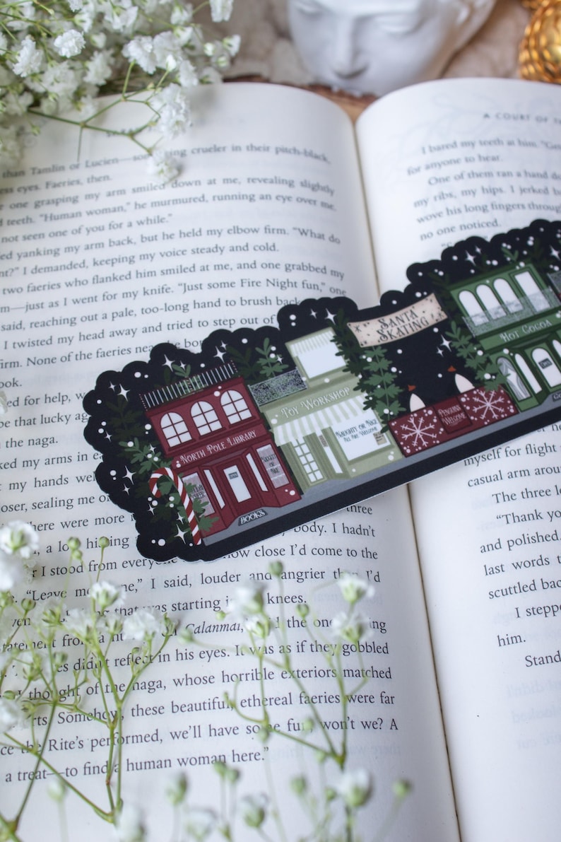 May include: A black and white illustrated sticker featuring a winter village scene with five buildings. The buildings are decorated with Christmas lights and snow. The buildings include a toy workshop, a library, a Santa skating rink, a hot cocoa shop, and a bakery. The sticker is perfect for adding a festive touch to any winter project.