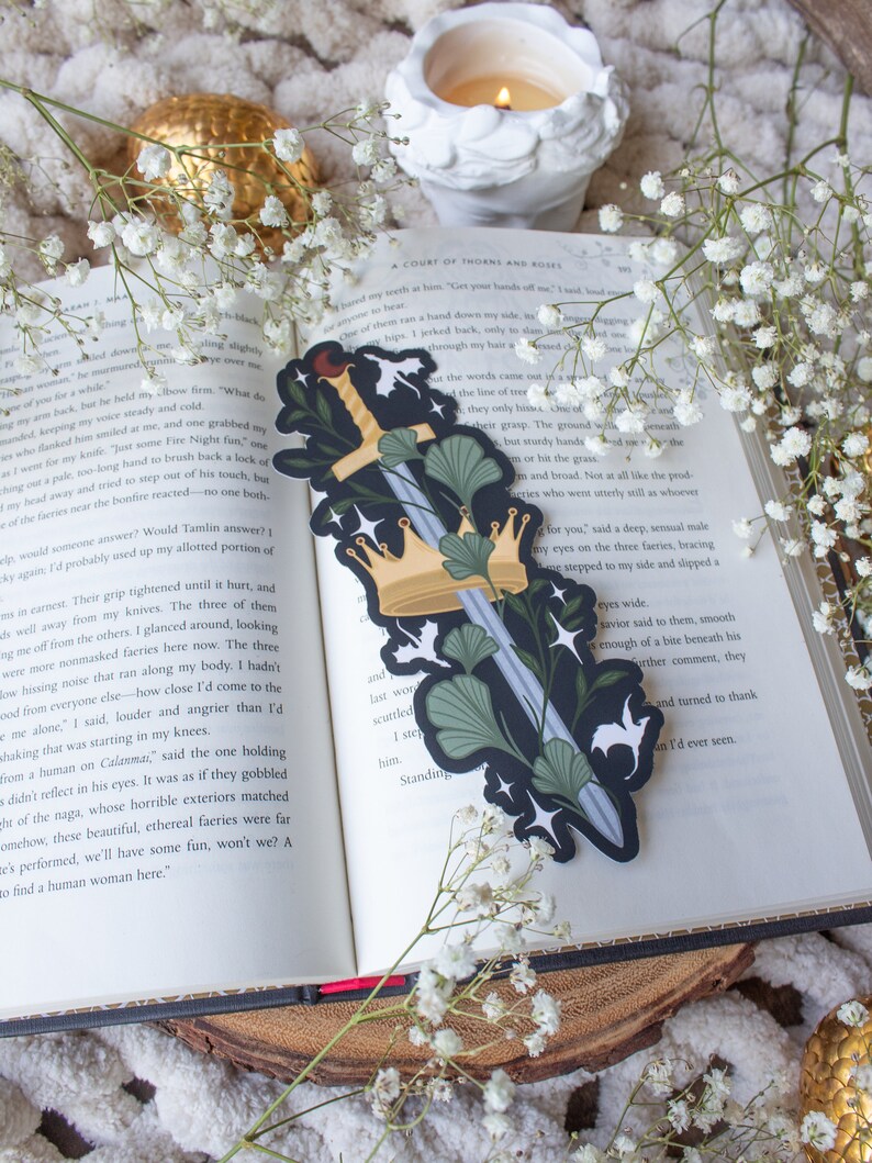 May include: A black and silver sword with a gold crown and green leaves. The sword is surrounded by white stars and a white background.