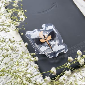 May include: A clear acrylic phone grip featuring a bat design with orange and black wings, a crescent moon, and clouds. The grip is placed on a dark gray e-reader, surrounded by white flowers and a book.