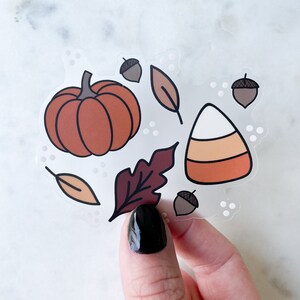 Puede incluir: Una hoja de pegatinas transparente con ilustraciones de tem&aacute;tica oto&ntilde;al. La hoja incluye una calabaza, un caramelo de ma&iacute;z, bellotas y hojas en tonos naranja, marr&oacute;n y rojo.
