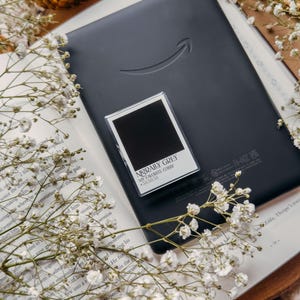 May include: A black e-reader with the Amazon logo rests on an open book, with a small, framed card that reads "MORALLY GREY MY FAVORITE COLOR 38.38.38." White flowers and gold decorative spheres surround the book.