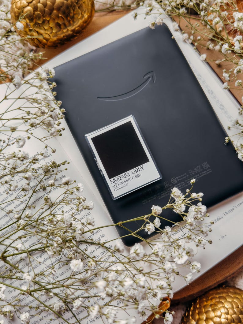 May include: A black e-reader with the Amazon logo rests on an open book, with a small, framed card that reads "MORALLY GREY MY FAVORITE COLOR 38.38.38." White flowers and gold decorative spheres surround the book.