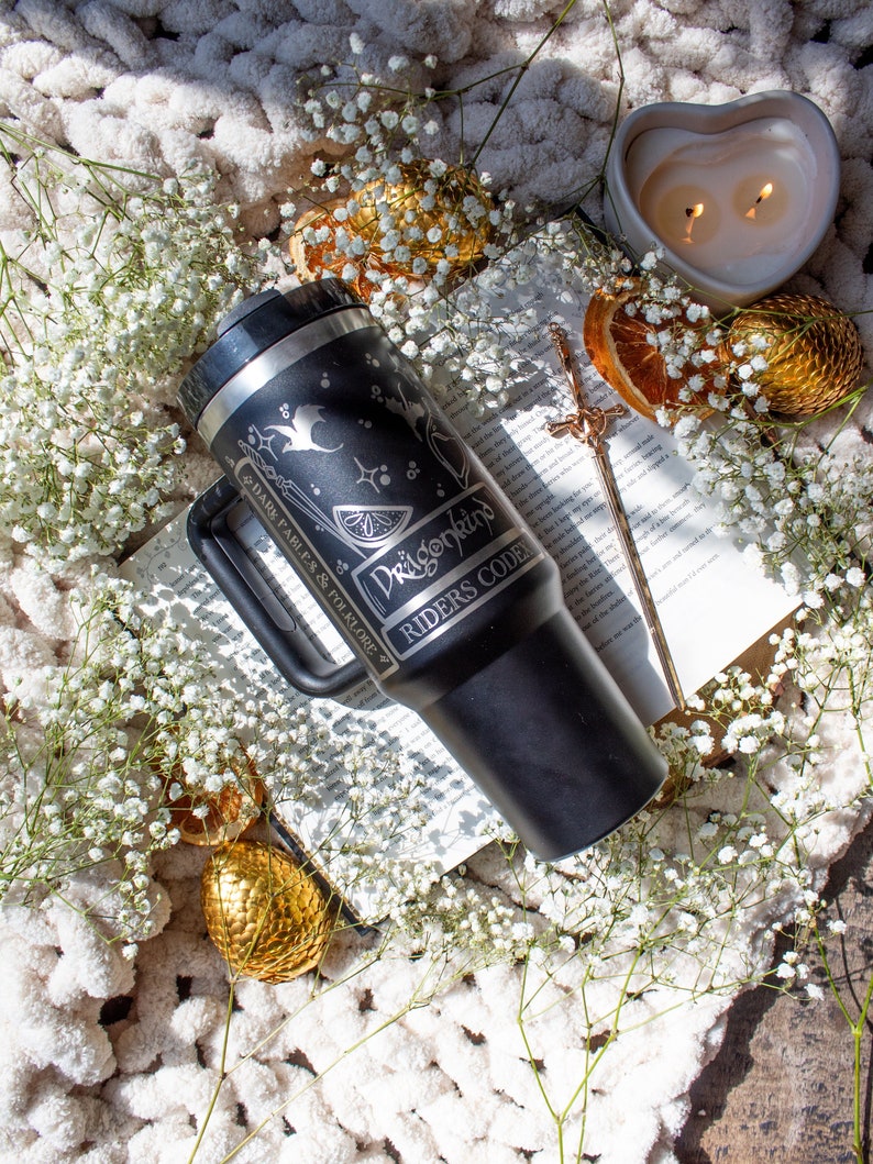May include: Black travel mug with a white design featuring a dragon, a lemon, and the text "Dragonkind Riders Code". The mug is sitting on a book with white flowers and a heart-shaped candle.