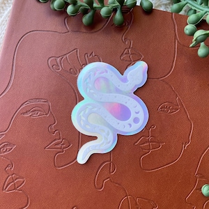 May include: A holographic sticker in the shape of a snake, with moon and leaf details, is placed on a brown leather-bound journal. The journal has embossed line art of faces. Green leafy sprigs are in the background.