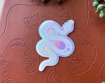May include: A holographic sticker in the shape of a snake, with moon and leaf details, is placed on a brown leather-bound journal. The journal has embossed line art of faces. Green leafy sprigs are in the background.