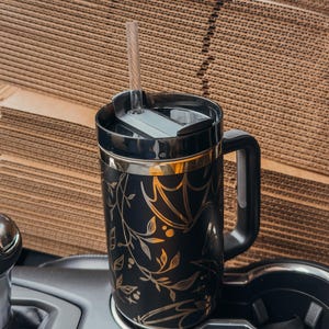 May include: Black travel mug with a gold floral and bat design. The mug has a black lid and a black handle. A clear straw is inserted into the lid.