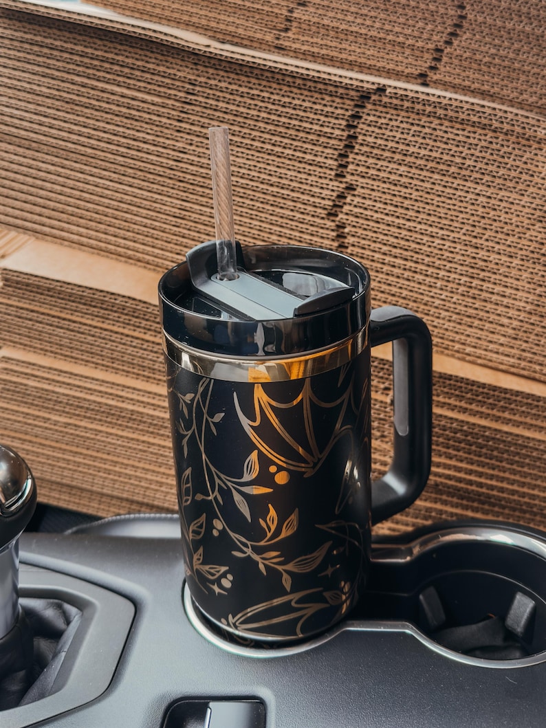 May include: Black travel mug with a gold floral and bat design. The mug has a black lid and a black handle. A clear straw is inserted into the lid.