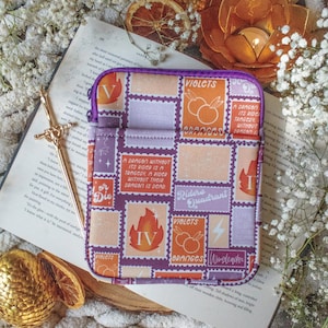 May include: A purple and orange tablet sleeve with a zipper closure. The sleeve features a stamp-like design with text including "Violets", "Oranges", and "A Dragon Without Its Rider Is A Tragedy". The sleeve is resting on an open book.