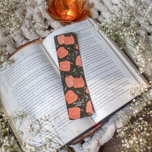 May include: A green and orange bookmark with a pumpkin design and the text "Meaggie Mood". The bookmark is lying on top of an open book with white flowers in the background.