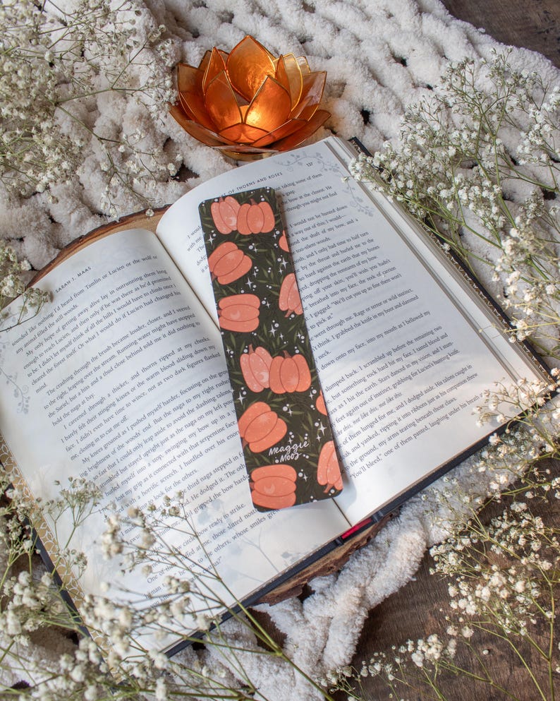 May include: A green and orange bookmark with a pumpkin design and the text "Meaggie Mood". The bookmark is lying on top of an open book with white flowers in the background.