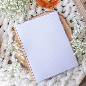 Puede incluir: Un cuaderno blanco con l&iacute;neas y una encuadernaci&oacute;n en espiral dorada sobre una manta blanca y esponjosa con un peque&ntilde;o portavelas naranja y flores blancas.