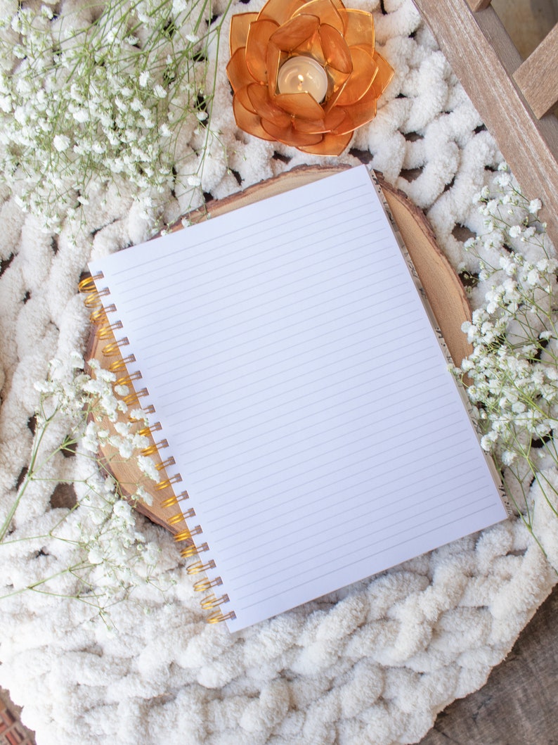 Puede incluir: Un cuaderno blanco con l&iacute;neas y una encuadernaci&oacute;n en espiral dorada sobre una manta blanca y esponjosa con un peque&ntilde;o portavelas naranja y flores blancas.