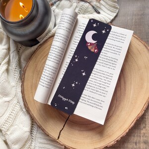 Puede incluir: Un marcap&aacute;ginas negro con una luna creciente, estrellas y una polilla con un patr&oacute;n rojo y amarillo. El marcap&aacute;ginas tiene el texto "Meaggie Moos" impreso en &eacute;l.