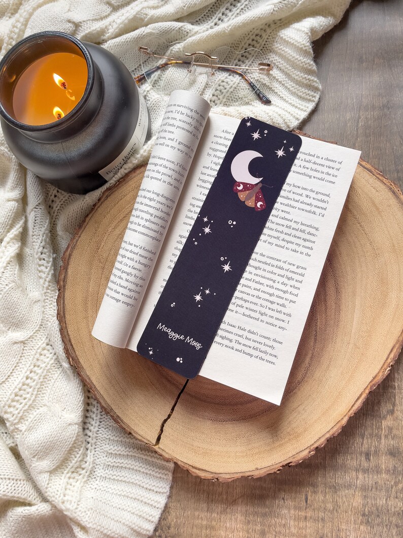 Puede incluir: Un marcap&aacute;ginas negro con una luna creciente, estrellas y una polilla con un patr&oacute;n rojo y amarillo. El marcap&aacute;ginas tiene el texto "Meaggie Moos" impreso en &eacute;l.