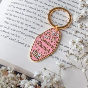 Puede incluir: Un llavero de esmalte rosa con borde dorado, con el texto "Romance Reader Book Club" y motivos florales y de corazón. El llavero está sobre un libro abierto, con flores blancas.