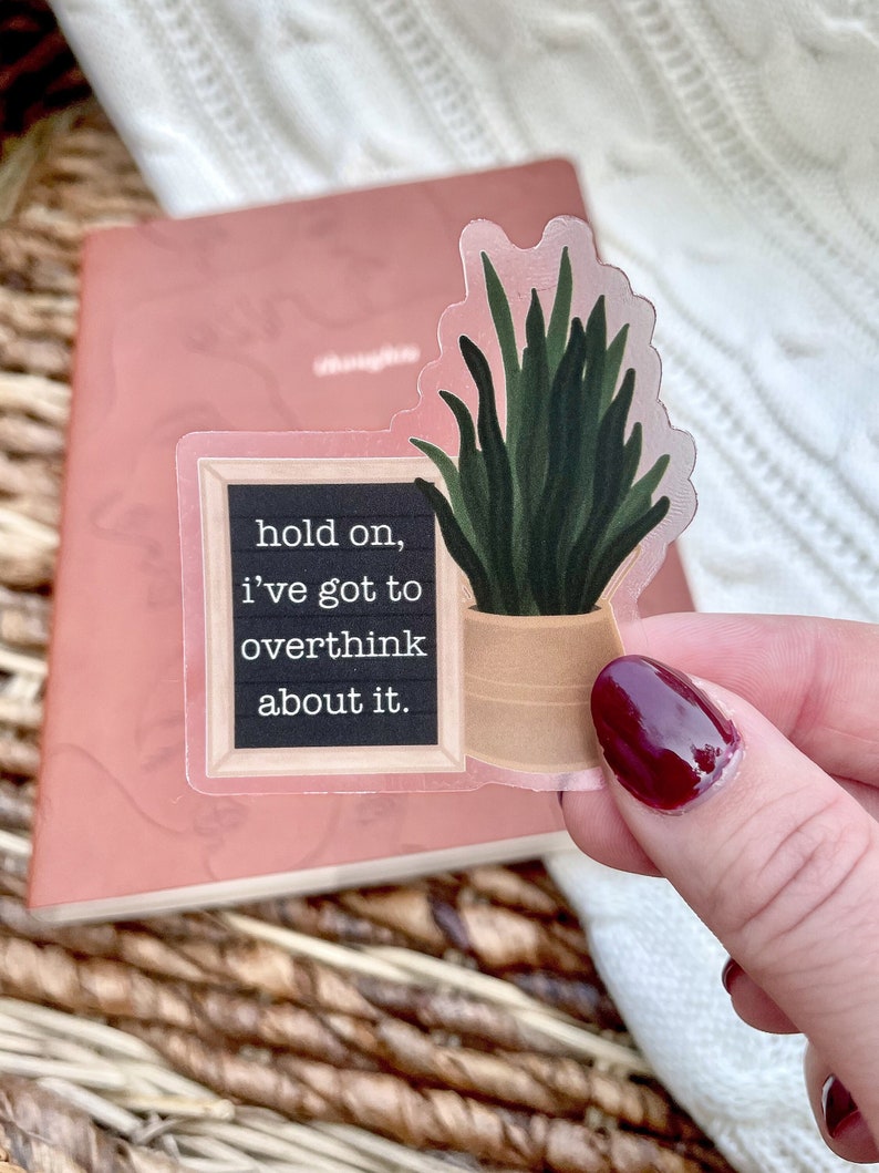 May include: A clear sticker with a black chalkboard background and white text that reads "hold on, i've got to overthink about it." A green plant in a brown pot is on the right side of the sticker.