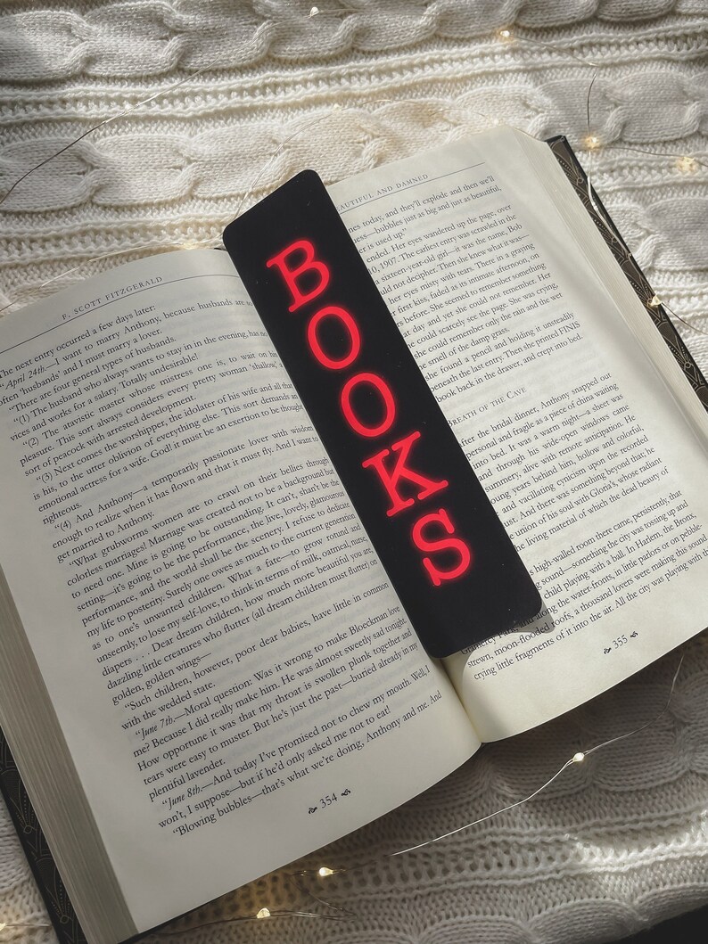May include: A close-up of a black bookmark with the word "BOOKS" in red neon letters. The bookmark is placed on an open book with white pages and black text. The book is titled "The Beautiful and Damned".