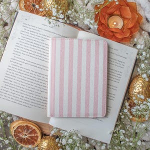 Puede incluir: Una cubierta de cuaderno a rayas rosas y blancas descansa sobre un libro abierto. La escena está decorada con rodajas de naranja secas, huevos decorativos dorados y un candelabro en forma de flor con una vela encendida. Flores blancas están dispersas.