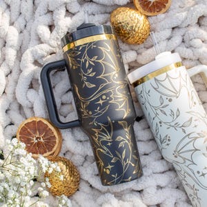 May include: Two stainless steel travel mugs with gold accents and a floral pattern. The mugs are on a white fluffy surface with dried citrus and gold ornaments.