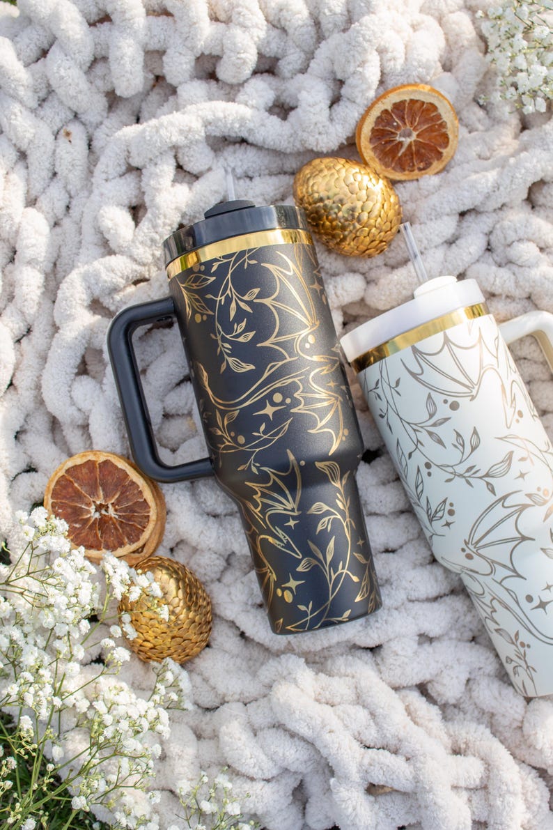 May include: Two stainless steel travel mugs with gold accents and a floral pattern. The mugs are on a white fluffy surface with dried citrus and gold ornaments.
