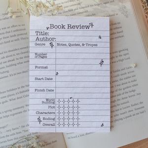 May include: A white Book Review card with a grid layout for notes, quotes, and tropes. The card has sections for title, author, genre, number of pages, format, start and finish dates, world building, plot, characters, ending, and overall. The card is surrounded by open books and flowers.