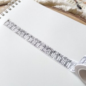 K&ouml;nnte beinhalten: Eine Rolle wei&szlig;es Washi-Klebeband mit einem schwarz-wei&szlig;en Muster aus gestapelten B&uuml;chern und floralen Akzenten.