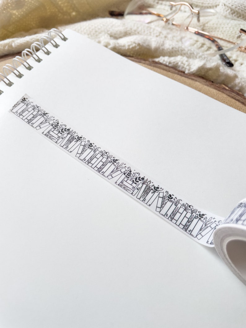 K&ouml;nnte beinhalten: Eine Rolle wei&szlig;es Washi-Klebeband mit einem schwarz-wei&szlig;en Muster aus gestapelten B&uuml;chern und floralen Akzenten.