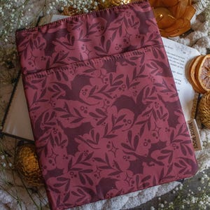 May include: A burgundy book sleeve with a repeating pattern of bats and holly leaves. The sleeve has a zipper closure and the text "Merch Moods" is printed on the bottom right corner.