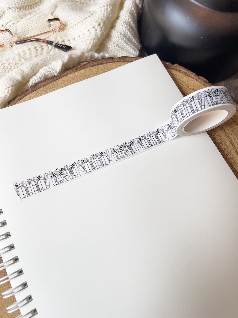 K&ouml;nnte beinhalten: Ein wei&szlig;es Washi-Tape mit einem schwarz-wei&szlig;en Muster aus B&uuml;chern und Blumen. Das Band ist aufgerollt und teilweise auf einem wei&szlig;en Notizbuch abgewickelt.