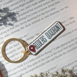 Op de afbeelding: Een witte emaille pin met een rood pin-icoon en de tekst "RIDERS QUADRANT" in zwarte letters. De pin is bevestigd aan een gouden sleutelhanger.