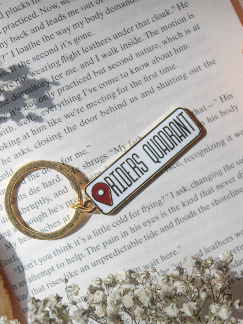 Op de afbeelding: Een witte emaille pin met een rood pin-icoon en de tekst "RIDERS QUADRANT" in zwarte letters. De pin is bevestigd aan een gouden sleutelhanger.