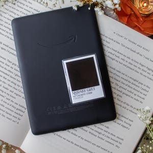 May include: A black e-reader with the Amazon logo, resting on an open book with white flowers. A small sticker with the text "MORALLY GREY MY FAVORITE COLOR #383838" is on the e-reader. The scene is lit with a warm, soft light.