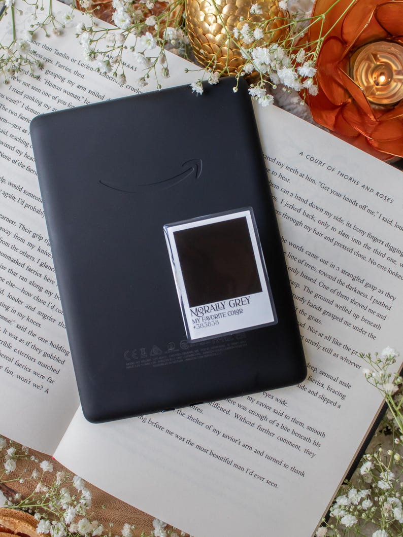 May include: A black e-reader with the Amazon logo, resting on an open book with white flowers. A small sticker with the text "MORALLY GREY MY FAVORITE COLOR #383838" is on the e-reader. The scene is lit with a warm, soft light.
