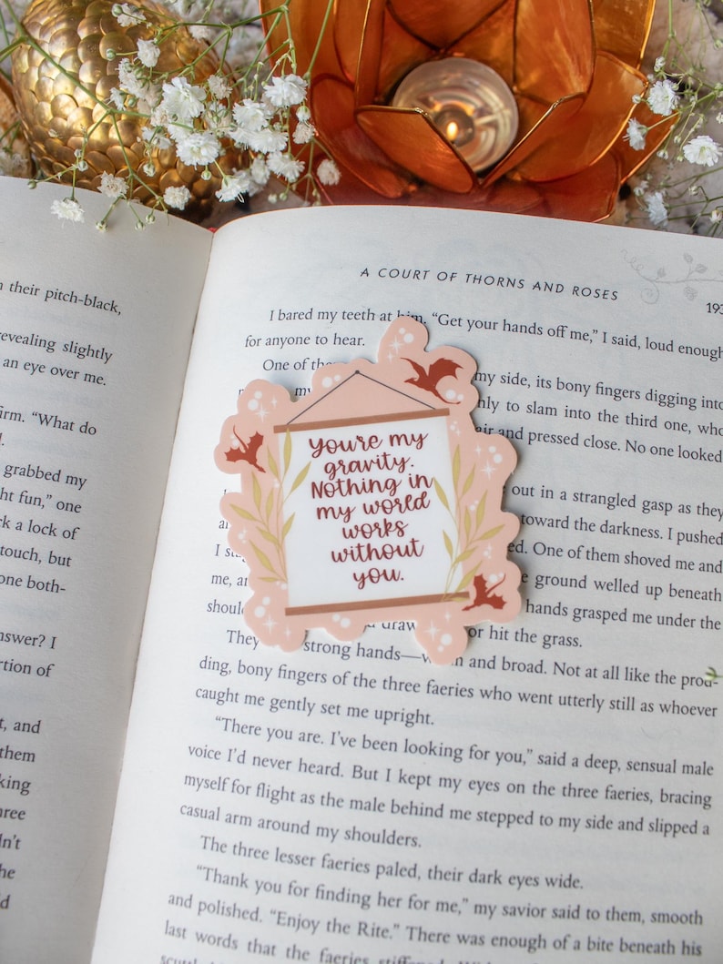 K&ouml;nnte beinhalten: Ein dekorativer Aufkleber mit einem rosa Rahmen und dem Text "you're my gravity. Nothing in my world works without you." Der Aufkleber hat ein florales Design und Fledermaus-Silhouetten. Er liegt auf einem offenen Buch.