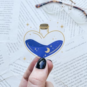 May include: A heart-shaped glass bottle with a blue liquid inside. The bottle is decorated with gold stars and a crescent moon. The bottle is on a white background.