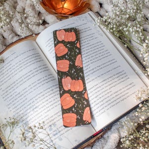 May include: A black bookmark with a repeating pattern of orange pumpkins and green leaves on a dark green background. The bookmark has a subtle shimmer effect and is accented with small white stars.