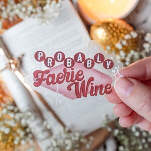 Puede incluir: Una pegatina transparente con el texto "Probably Faerie Wine" en letras rosa y blancas. La pegatina está sobre un fondo blanco con un libro y flores blancas.