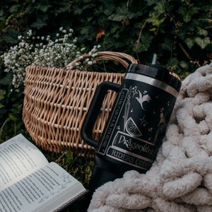 May include: A black and silver stainless steel tumbler with a dragon design and the text "Dragonkind Riders Code" and "Dark Fables & Folklore". The tumbler is sitting on a white knitted blanket next to a wicker basket and an open book.