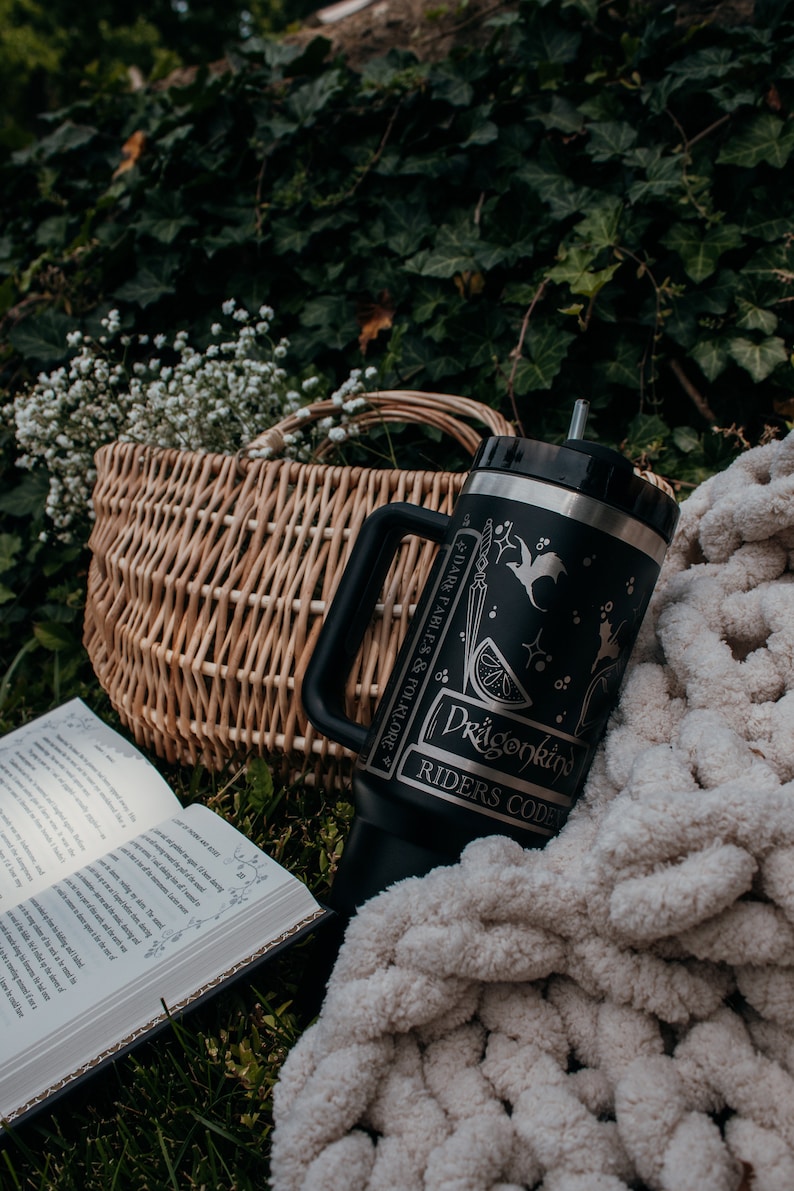 May include: A black and silver stainless steel tumbler with a dragon design and the text "Dragonkind Riders Code" and "Dark Fables & Folklore". The tumbler is sitting on a white knitted blanket next to a wicker basket and an open book.